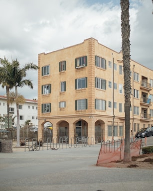 A four-story, beige apartment building with numerous windows and arched entrances stands prominently. The foreground features bike racks and palm trees, with a cloudy sky overhead.
