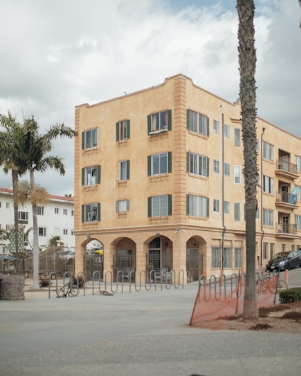 A four-story, beige apartment building with numerous windows and arched entrances stands prominently. The foreground features bike racks and palm trees, with a cloudy sky overhead.