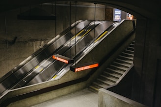 An underground subway station featuring a modern escalator and a set of stairs. The escalator is flanked by a concrete wall and is illuminated by overhead lights. Signage indicates directions to Montmorency and Cote-Vertu. The environment gives a sense of industrial architecture with a hint of minimalism.