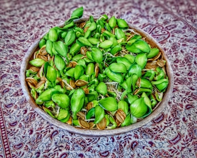 A wooden bowl filled with green sprouting seeds and brown seeds lies on a patterned fabric with intricate designs in shades of red and gray.