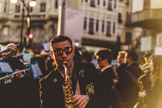 A musician wearing sunglasses plays a saxophone in a bustling street in front of a crowd, as part of a marching band. Several other musicians are visible in the background holding instruments, and sheet music is attached to some of the instruments. The atmosphere is lively with warm, golden sunlight casting a glow over the scene. Tall buildings line the street and pedestrians can be seen in the blurred background.