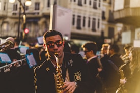 A musician wearing sunglasses plays a saxophone in a bustling street in front of a crowd, as part of a marching band. Several other musicians are visible in the background holding instruments, and sheet music is attached to some of the instruments. The atmosphere is lively with warm, golden sunlight casting a glow over the scene. Tall buildings line the street and pedestrians can be seen in the blurred background.