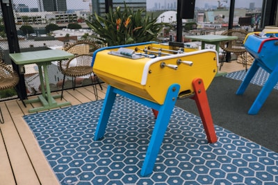 A foosball table with vibrant colors stands on a patterned blue mat. The structure features a yellow body with red and blue legs, and silver rods with handles are visible. Surrounding the table are wicker chairs and green square tables, set on a rooftop or terrace area. The background includes a cityscape with buildings and greenery.