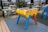 A foosball table with vibrant colors stands on a patterned blue mat. The structure features a yellow body with red and blue legs, and silver rods with handles are visible. Surrounding the table are wicker chairs and green square tables, set on a rooftop or terrace area. The background includes a cityscape with buildings and greenery.