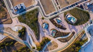 An aerial view of an abstractly designed park featuring winding pathways, geometric playground structures, and landscaped areas with grass and trees. The park is surrounded by roads and urban development, and there are recreational spaces with some people visible.