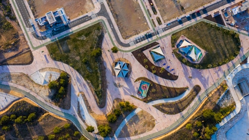 An aerial view of an abstractly designed park featuring winding pathways, geometric playground structures, and landscaped areas with grass and trees. The park is surrounded by roads and urban development, and there are recreational spaces with some people visible.