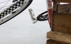 A close-up view of a bicycle showing the rear wheel with a checkered pattern and a pedal visible near a wooden pallet. The frame is red with part of a chain and gear mechanism visible.