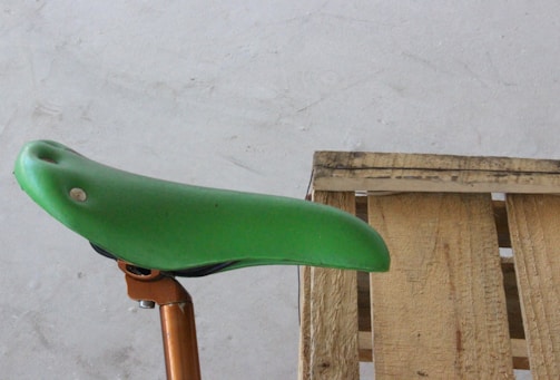 A green bicycle seat is mounted on an orange seat post next to a wooden pallet on a concrete floor. The image focuses on the contrast between the smooth, colorful surface of the seat and the rough texture of the pallet.