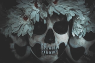 A close-up view of three intricately painted skull masks, adorned with white flowers forming a crown. The masks display detailed black and white patterns reminiscent of Day of the Dead (Día de los Muertos) face paint. The flowers add a contrasting softness to the otherwise dark and eerie skull imagery.