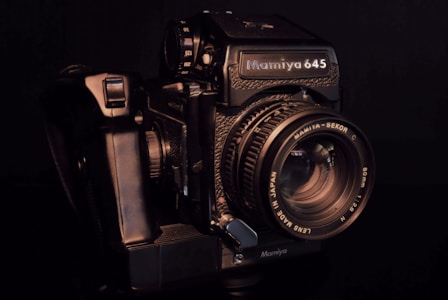 A vintage Mamiya 645 medium format camera is prominently displayed, featuring a detailed lens with markings indicating specifications. The camera's body is black with textured grips and metallic components, evoking a sense of nostalgia and precision craftsmanship.