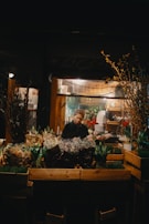 A flower shop at night with a person standing behind a display of various bouquets and plants. Soft lighting from inside the shop illuminates the scene, highlighting the colorful array of flowers, including tulips and hyacinths. Wooden crates are used to display the plants and the background suggests a cozy, inviting space.