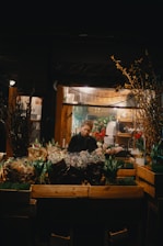 A flower shop at night with a person standing behind a display of various bouquets and plants. Soft lighting from inside the shop illuminates the scene, highlighting the colorful array of flowers, including tulips and hyacinths. Wooden crates are used to display the plants and the background suggests a cozy, inviting space.