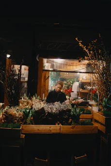 A flower shop at night with a person standing behind a display of various bouquets and plants. Soft lighting from inside the shop illuminates the scene, highlighting the colorful array of flowers, including tulips and hyacinths. Wooden crates are used to display the plants and the background suggests a cozy, inviting space.
