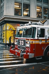 A red fire truck with the number 33 and the New York City Fire Department insignia is parked on a wet city street. It is a rainy day, and the street is shiny from the moisture. In the background, there is a building with large glass windows and a person walking under an umbrella.
