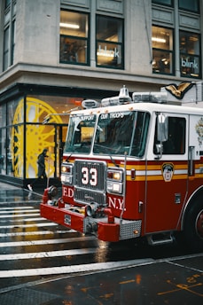 A red fire truck with the number 33 and the New York City Fire Department insignia is parked on a wet city street. It is a rainy day, and the street is shiny from the moisture. In the background, there is a building with large glass windows and a person walking under an umbrella.