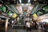 A busy urban escalator and stairway area features vibrant advertisements for a telecommunications company on both sides, with numerous people ascending and descending. The setting appears to be indoors, possibly a transit station, with a ceiling lined with fluorescent lights.