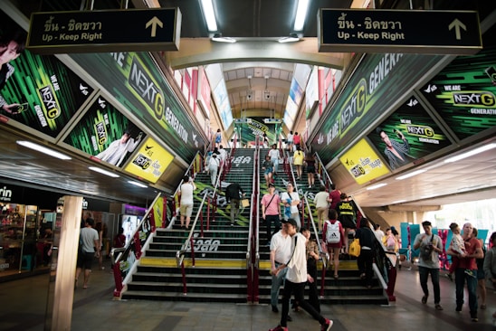 A busy urban escalator and stairway area features vibrant advertisements for a telecommunications company on both sides, with numerous people ascending and descending. The setting appears to be indoors, possibly a transit station, with a ceiling lined with fluorescent lights.