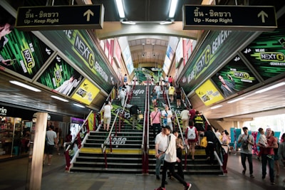 A busy urban escalator and stairway area features vibrant advertisements for a telecommunications company on both sides, with numerous people ascending and descending. The setting appears to be indoors, possibly a transit station, with a ceiling lined with fluorescent lights.