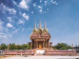 A richly decorated temple stands majestically under a vibrant blue sky. The architecture features intricate patterns and ornate spires, surrounded by an open paved area with some greenery in the background. The temple displays a blend of gold, red, and green hues, reflecting traditional Southeast Asian design elements.