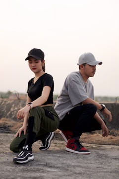A woman and a man are squatting on a concrete surface, both wearing casual streetwear clothing. The woman is dressed in a black cap, black t-shirt, olive green pants, and black sneakers, while the man wears a gray cap, gray t-shirt, dark pants, and red sneakers. The background is a muted outdoor setting with overcast lighting.