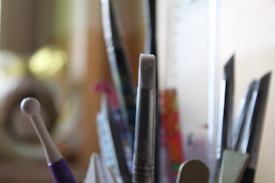 Various art supplies, including paintbrushes and other tools, are placed in a container. The focus is on the tip of a white brush, with other tools out of focus in the background.