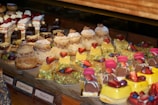 A display of assorted pastries and desserts including eclairs, layered cakes, fruit tarts, and small cakes topped with strawberries, chocolate, and cream. The pastries are arranged on a wooden counter, showcasing a variety of textures and shapes, with a focus on vibrant toppings such as pistachios and berries.