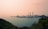 A coastal industrial facility features multiple tall chimneys and utility structures situated by the sea. The foreground includes lush green vegetation on a hill contrasting with the industrial area. The sky is hazy with a pinkish hue, suggesting pollution or sunrise/sunset conditions.