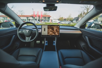 The interior of an electric car featuring a minimalist dashboard design with a central touchscreen display. The steering wheel prominently displays the car company's logo. Leather seats and a sleek wooden trim enhance the modern aesthetic. Outside the car, a backdrop of buildings and parked vehicles is visible through the windshield.