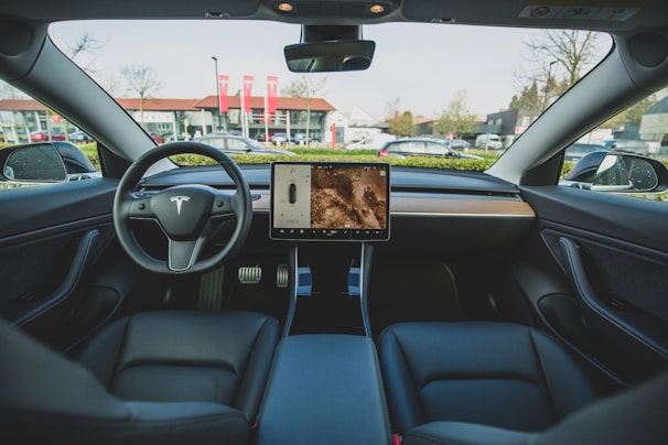 The interior of an electric car featuring a minimalist dashboard design with a central touchscreen display. The steering wheel prominently displays the car company's logo. Leather seats and a sleek wooden trim enhance the modern aesthetic. Outside the car, a backdrop of buildings and parked vehicles is visible through the windshield.