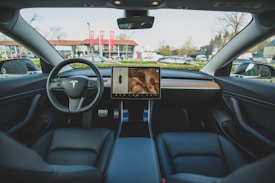 The interior of an electric car featuring a minimalist dashboard design with a central touchscreen display. The steering wheel prominently displays the car company's logo. Leather seats and a sleek wooden trim enhance the modern aesthetic. Outside the car, a backdrop of buildings and parked vehicles is visible through the windshield.