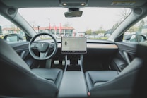 The interior of a modern car with a minimalistic design. A large touchscreen display is centrally mounted on the dashboard, displaying a map. The steering wheel features a logo with a distinctive letter design. The seats and dashboard are made of sleek, dark materials with hints of wood trim. Large windows provide a view of a parking lot outside.