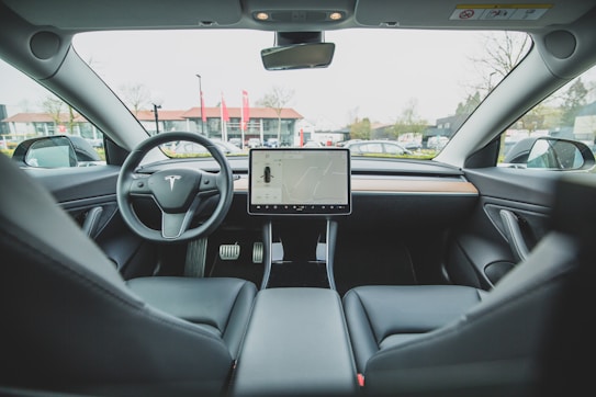 The interior of a modern car with a minimalistic design. A large touchscreen display is centrally mounted on the dashboard, displaying a map. The steering wheel features a logo with a distinctive letter design. The seats and dashboard are made of sleek, dark materials with hints of wood trim. Large windows provide a view of a parking lot outside.