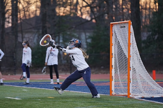 A lacrosse goalie is in motion, reaching out with a stick to catch or block a ball in front of the goal. Other players in uniforms and goggles are visible in the background on a sports field, with a backdrop of trees and a setting sun.