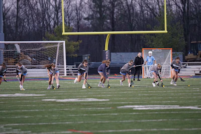 A group of young athletes are practicing on a sports field, holding lacrosse sticks and running in unison. A coach stands nearby, and a goalie is visible near the goalpost, which is surrounded by a net. The field shows yard marker lines and a large goalpost looms in the background. It's dusk, with trees visible in the distance.