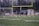A group of young athletes are practicing on a sports field, holding lacrosse sticks and running in unison. A coach stands nearby, and a goalie is visible near the goalpost, which is surrounded by a net. The field shows yard marker lines and a large goalpost looms in the background. It's dusk, with trees visible in the distance.