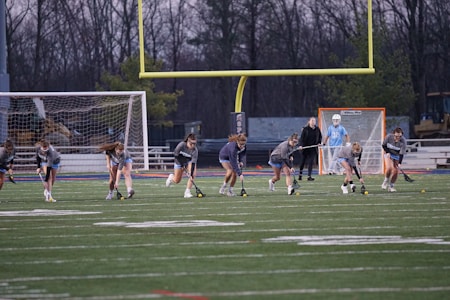 A group of young athletes are practicing on a sports field, holding lacrosse sticks and running in unison. A coach stands nearby, and a goalie is visible near the goalpost, which is surrounded by a net. The field shows yard marker lines and a large goalpost looms in the background. It's dusk, with trees visible in the distance.