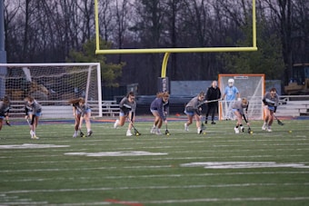 A group of young athletes are practicing on a sports field, holding lacrosse sticks and running in unison. A coach stands nearby, and a goalie is visible near the goalpost, which is surrounded by a net. The field shows yard marker lines and a large goalpost looms in the background. It's dusk, with trees visible in the distance.