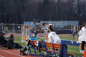 A group of lacrosse players gathers near the sideline, with several holding up their sticks in a team huddle. They are wearing matching uniforms with shorts and grey tops. Some people are seated on the benches nearby, possibly coaches or teammates. The field is lined and surrounded by a running track, with trees and a construction area visible in the background.