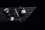 A set of grooming tools and products including a shaving brush, a razor, a bottle of healing post shave spray, and a jar of ultra lather shaving cream arranged on a dark tiled surface with a pattern of hexagonal tiles. The image features dramatic lighting with deep shadows.