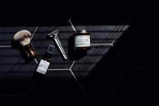 A set of grooming tools and products including a shaving brush, a razor, a bottle of healing post shave spray, and a jar of ultra lather shaving cream arranged on a dark tiled surface with a pattern of hexagonal tiles. The image features dramatic lighting with deep shadows.