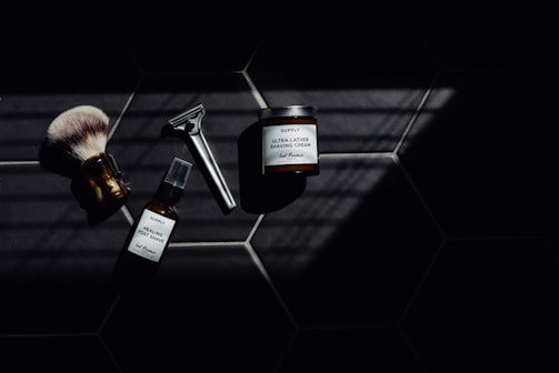 A set of grooming tools and products including a shaving brush, a razor, a bottle of healing post shave spray, and a jar of ultra lather shaving cream arranged on a dark tiled surface with a pattern of hexagonal tiles. The image features dramatic lighting with deep shadows.