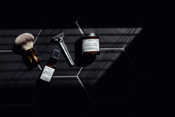 A set of grooming tools and products including a shaving brush, a razor, a bottle of healing post shave spray, and a jar of ultra lather shaving cream arranged on a dark tiled surface with a pattern of hexagonal tiles. The image features dramatic lighting with deep shadows.