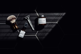 A set of grooming tools and products including a shaving brush, a razor, a bottle of healing post shave spray, and a jar of ultra lather shaving cream arranged on a dark tiled surface with a pattern of hexagonal tiles. The image features dramatic lighting with deep shadows.