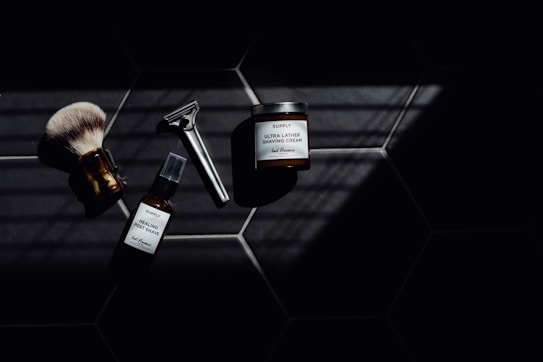 A set of grooming tools and products including a shaving brush, a razor, a bottle of healing post shave spray, and a jar of ultra lather shaving cream arranged on a dark tiled surface with a pattern of hexagonal tiles. The image features dramatic lighting with deep shadows.