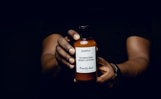 Two hands holding a bottle of hydrating body lotion against a dark background. The label is prominently visible, highlighting the brand and product information.