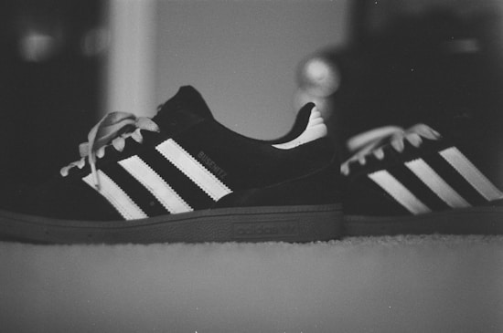 A pair of Adidas sneakers with the classic three-stripe design is captured in a monochrome setting. The sneakers are positioned on a soft surface with laces untied, showcasing their casual style.