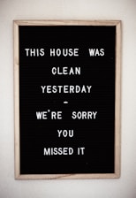A framed black letter board with white letters spelling out a humorous message about the house being clean yesterday. The sign is simple with plastic letters arranged in a casual style.