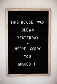 A framed black letter board with white letters spelling out a humorous message about the house being clean yesterday. The sign is simple with plastic letters arranged in a casual style.