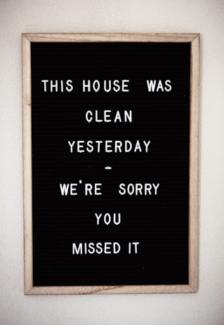 A framed black letter board with white letters spelling out a humorous message about the house being clean yesterday. The sign is simple with plastic letters arranged in a casual style.