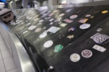 A luggage conveyor belt is covered with various colorful stickers. The surface is metallic and black, providing contrast to the stickers. Each sticker has unique designs and logos, and a piece of luggage is also partially visible on the conveyor.
